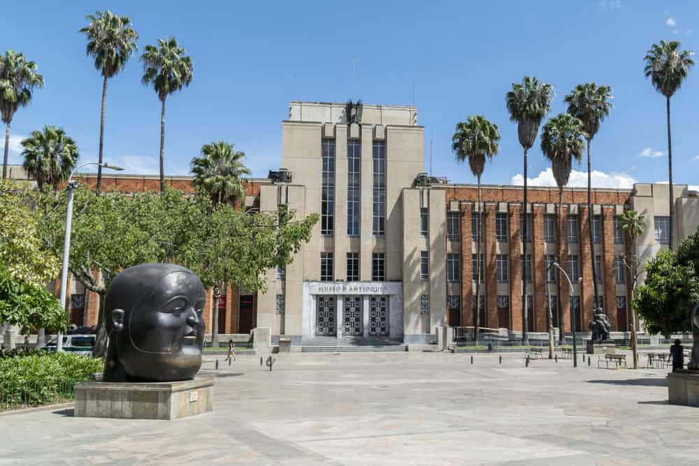 A plaza with palm trees, a large head sculpture, and a tall building labeled “Museo de Antioquia” offers one of the top Things to Do in Medellin with Kids, providing an engaging outdoor space and captivating art for families to explore together.
