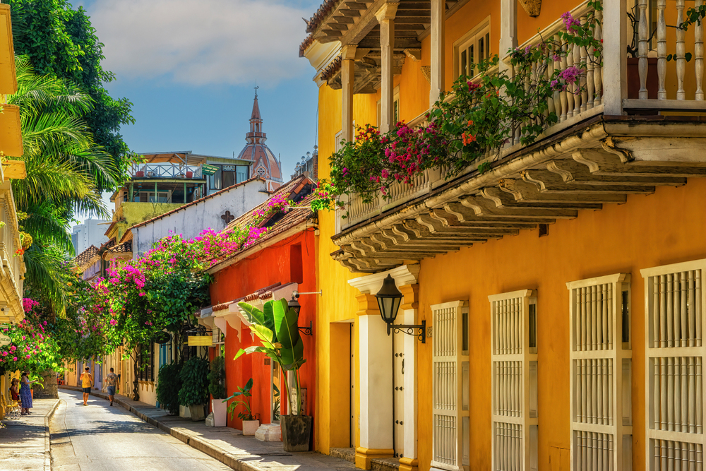 Colorful colonial buildings with flower-filled balconies line a sunlit, narrow street in Cartagena, Colombia, offering a charming glimpse of the vibrant city and serving as the perfect starting point for discovering the many things to do in Cartagena.