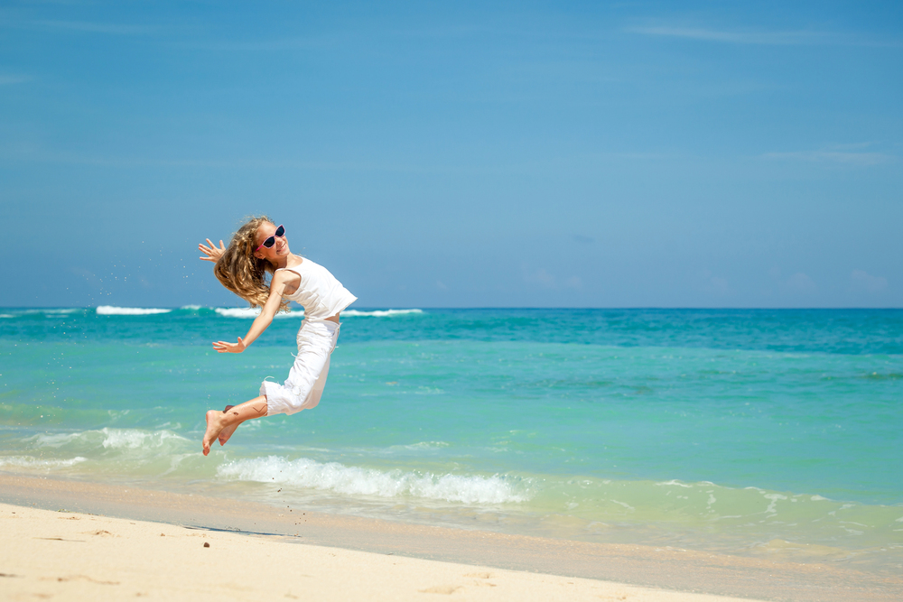 Woman in white clothes jumps joyfully on a sandy beach by clear turquoise water under a blue sky, embodying the perfect carefree moment during a travel with teens adventure.