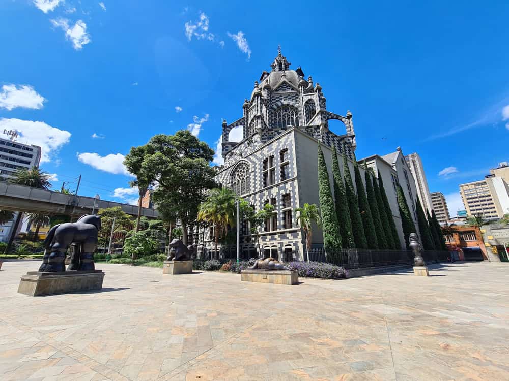 Gothic-style building with statues and trees in a sunny, open plaza under a bright blue sky—an inviting spot to explore as part of your list of things to do in Medellin with kids.