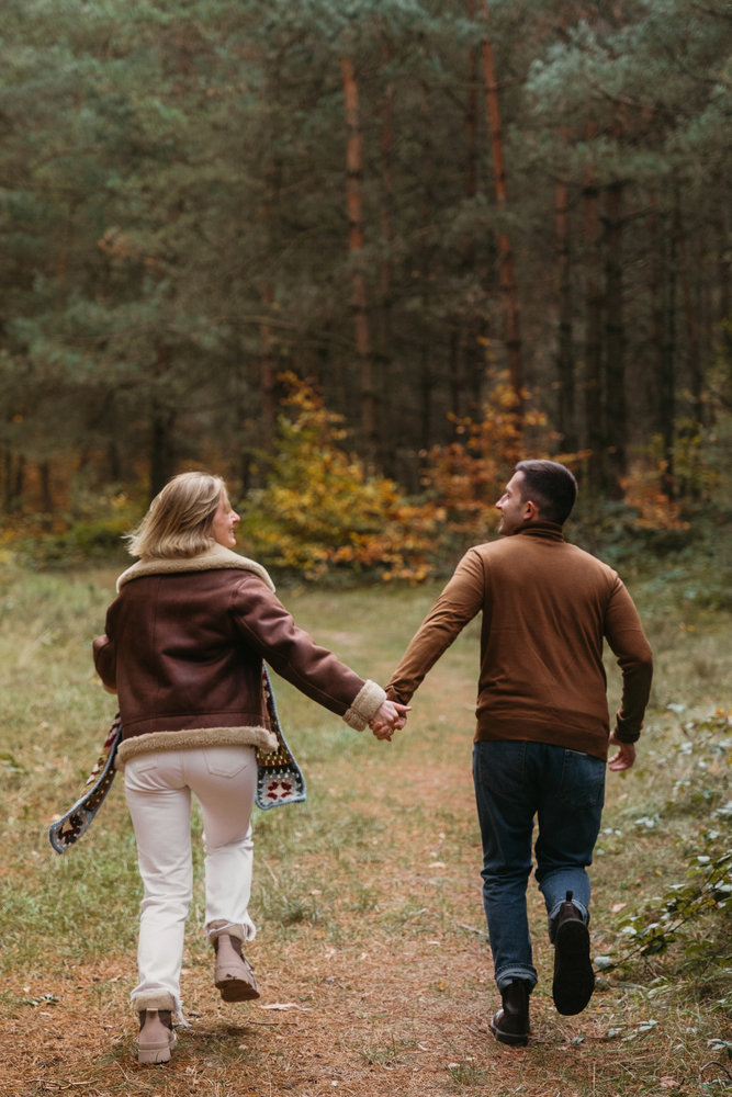A couple holding hands, walking on a forest path, surrounded by trees and autumn foliage&mdash;an inspiring scene of empty nest travel as they embark on new adventures together.