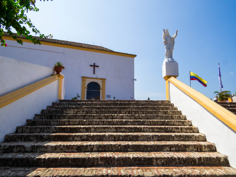 Stone steps lead to a white church with a cross, statue, and two flags under a clear blue sky&mdash;one of the many remarkable sights among the things to do in Cartagena.