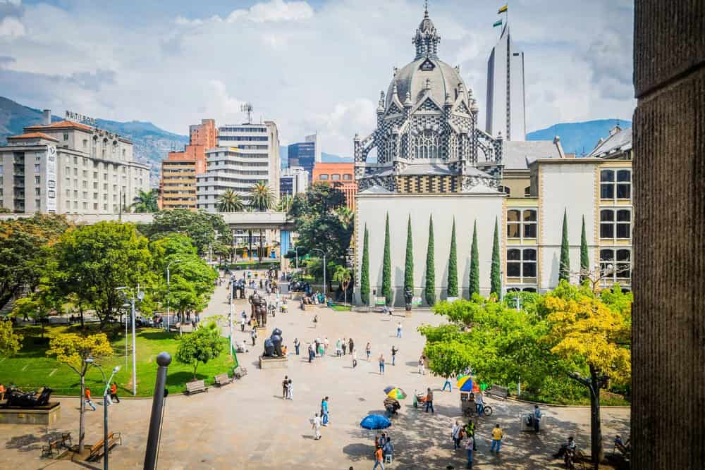 People walk in a sunny city plaza with trees and a historic building with domes in the background—an inviting spot perfect for families seeking things to do in Medellin with kids.