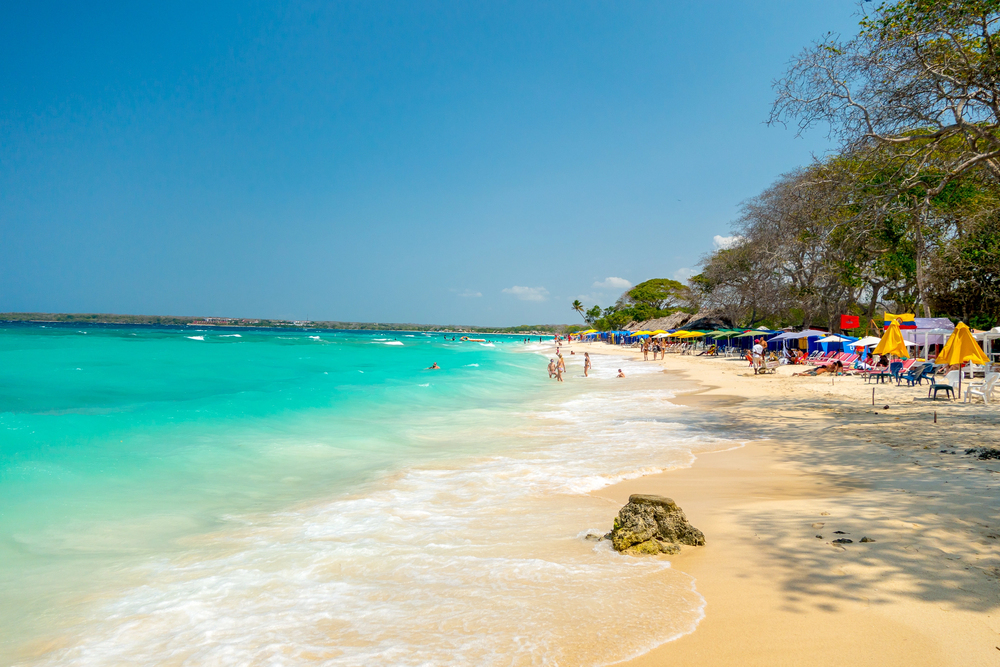 Sandy beach with turquoise water, people swimming, and sun umbrellas along the shoreline under a clear sky&mdash;one of the top things to do in Cartagena for a relaxing day by the sea.