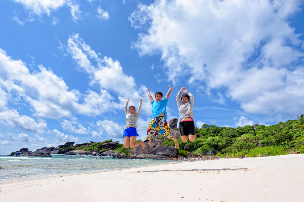 Three people jump in the air on a sandy beach with blue sky, clouds, and green trees in the background, capturing a joyful moment of travel with teens.