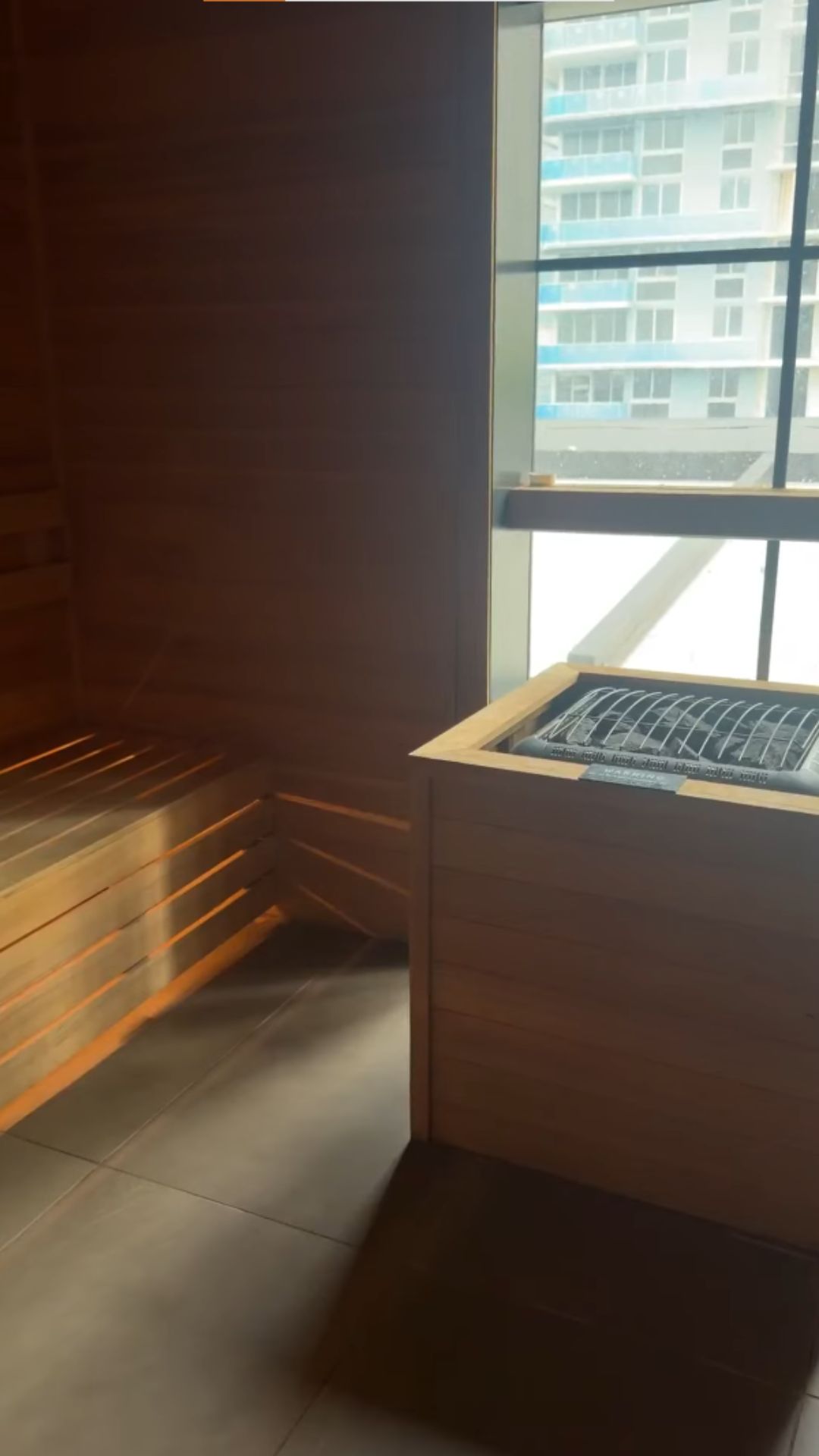 Sunlit wooden sauna room with bench seating and a heater, next to a large window overlooking buildings—an inviting retreat reminiscent of the serene wellness spaces highlighted in the Gale Miami Hotel & Residences Review.