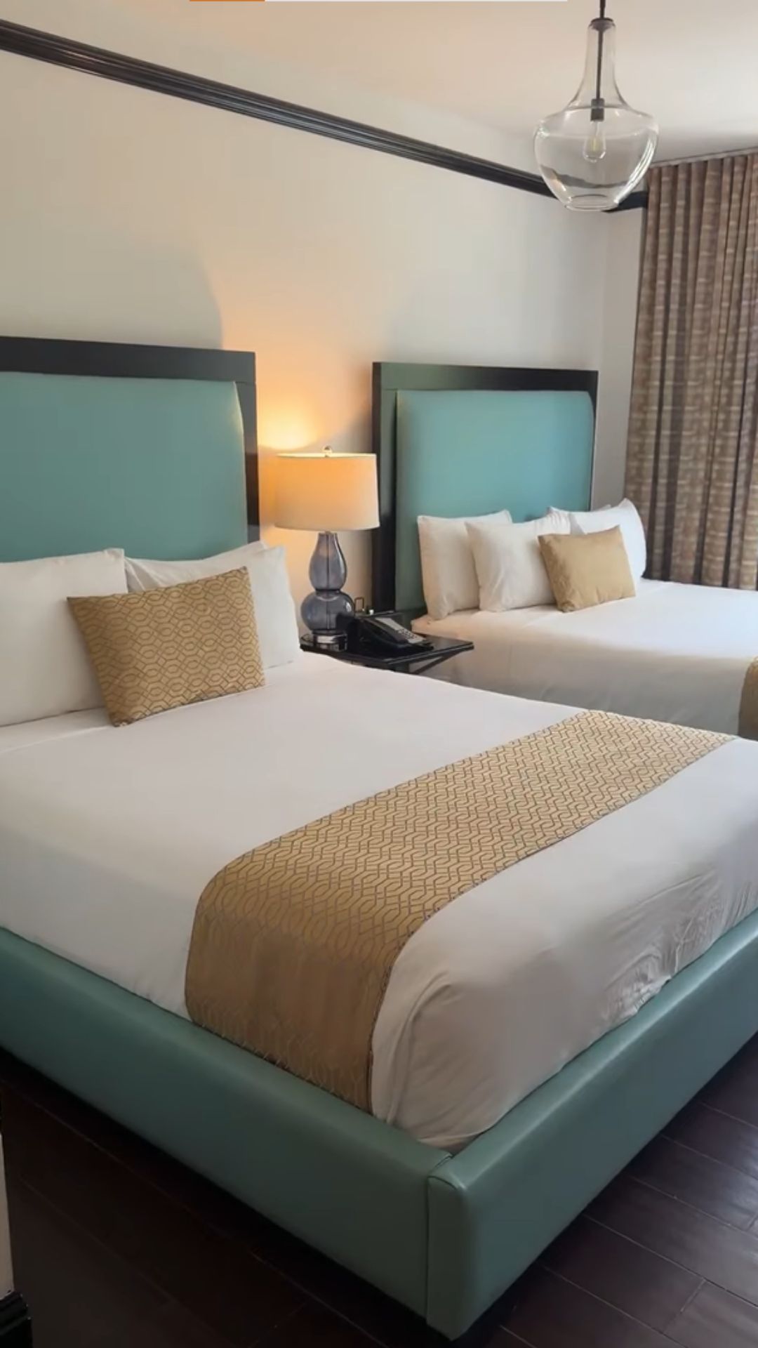 Hotel room with two teal beds, white bedding, gold pillows, a lamp, and beige curtains—perfect for travelers seeking comfort and style as mentioned in many Gale Miami Hotel & Residences reviews.