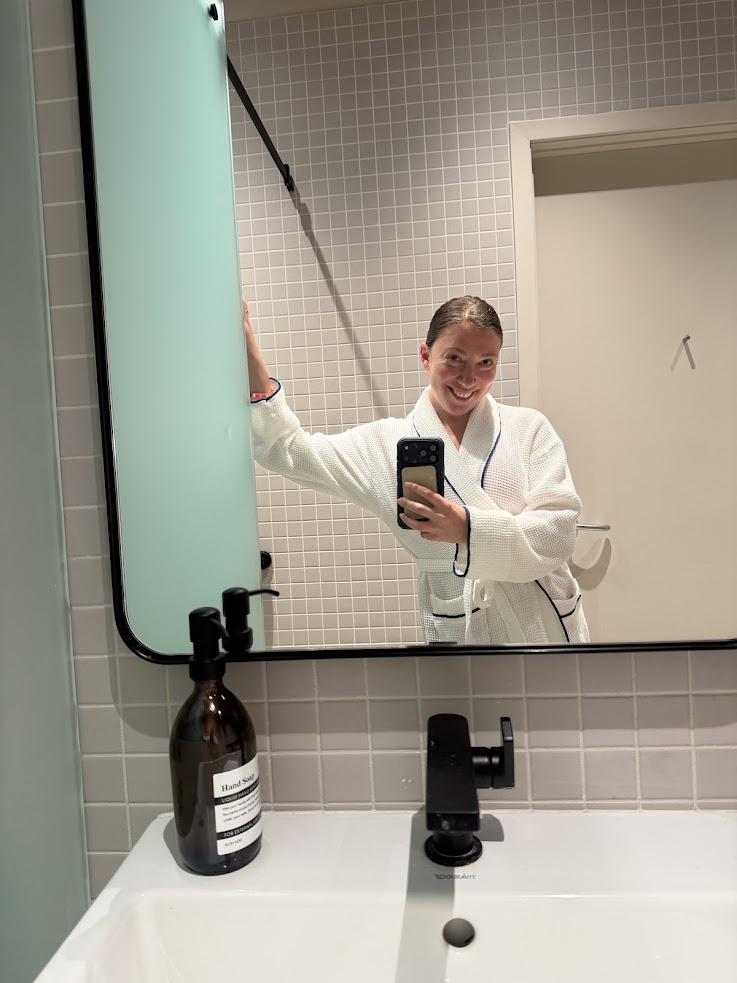 A person in a white robe takes a mirror selfie in a modern bathroom with tiled walls and a black faucet, capturing the chic ambiance reminiscent of what you’d find in a Gale Miami Hotel & Residences Review.