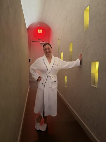 A person in a white robe and slippers stands smiling in a hallway with yellow-lit wall niches, capturing the relaxed ambiance highlighted in any Gale Miami Hotel & Residences Review.