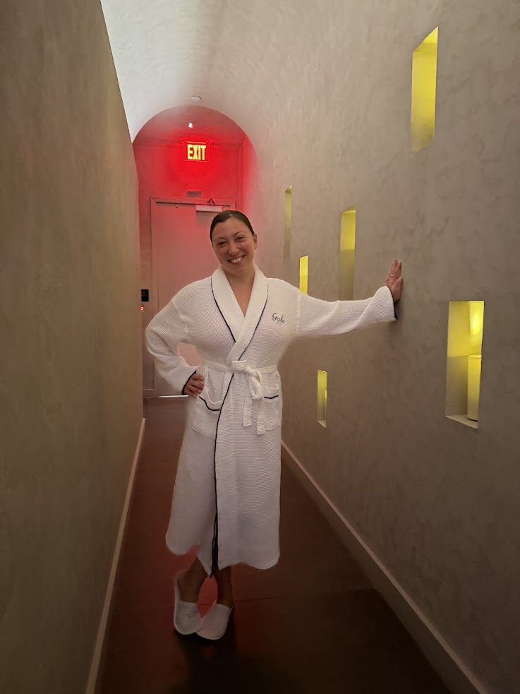 A person in a white robe and slippers stands smiling in a hallway with yellow-lit wall niches, capturing the relaxed ambiance highlighted in any Gale Miami Hotel & Residences Review.
