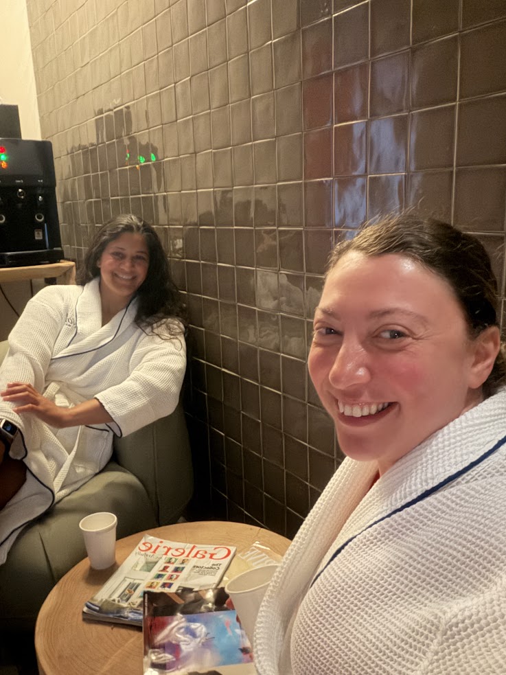Two women in white robes relax in a spa lounge, smiling, with magazines and drinks on a small table—a scene that perfectly captures the luxurious ambiance described in many Gale Miami Hotel & Residences reviews.