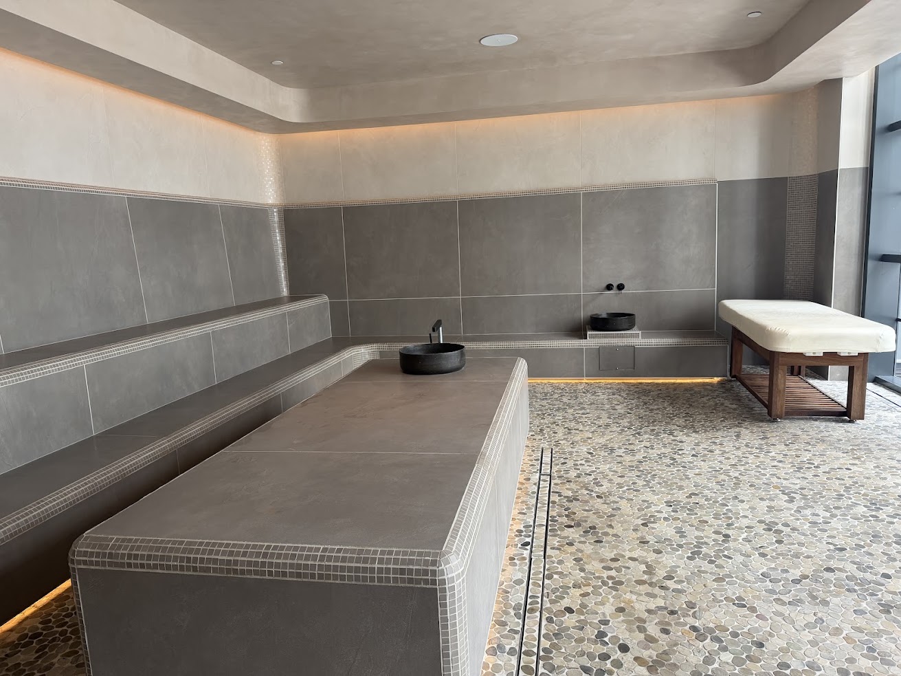 Modern spa room with tiled benches, a stone basin, and a cushioned massage table on a pebble-patterned floor—an atmosphere reminiscent of the tranquil wellness spaces highlighted in many Gale Miami Hotel & Residences reviews.