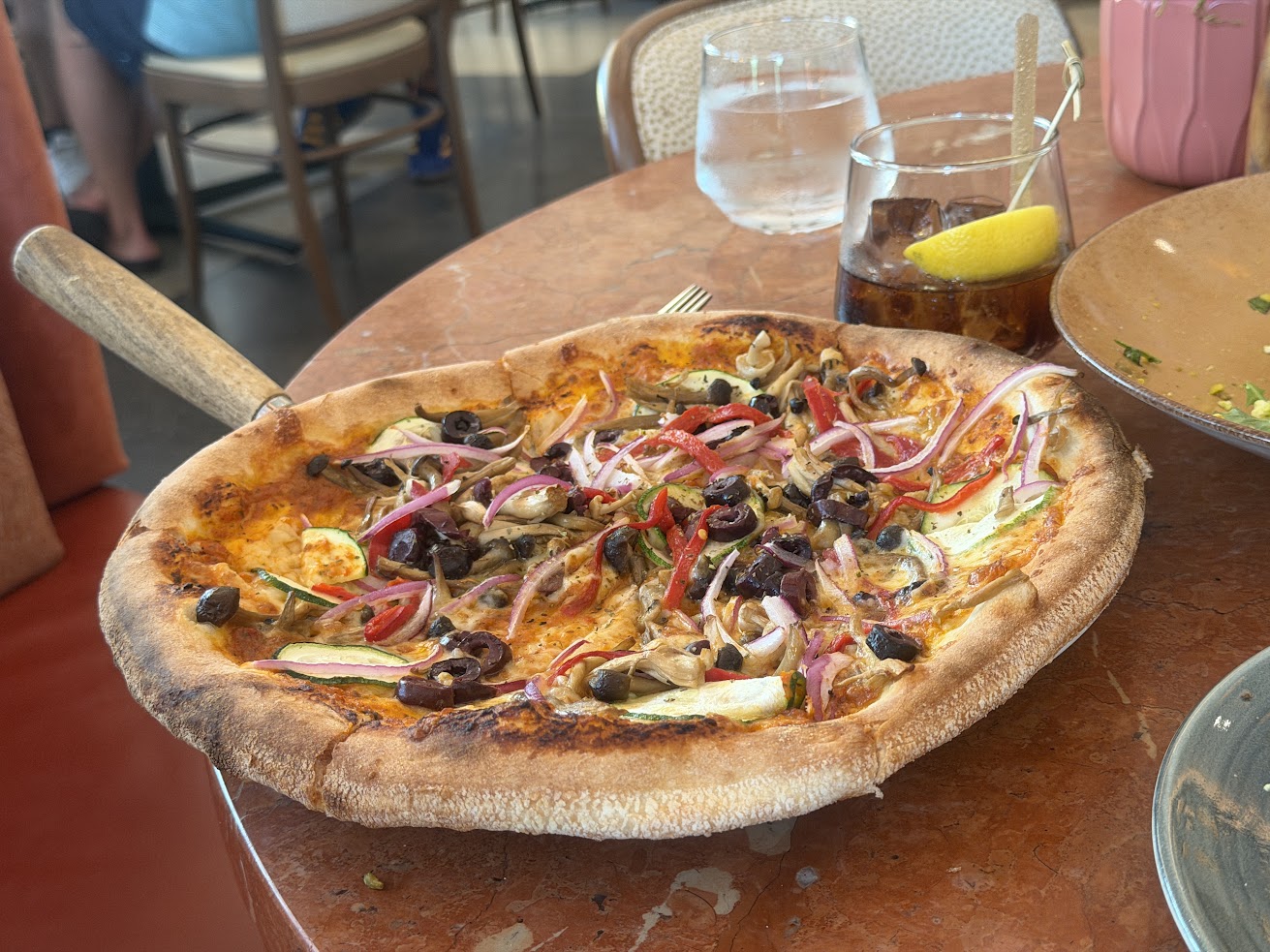 Wood-fired vegetable pizza with olives, red onions, and peppers on a table with drinks and a plate—an inviting scene reminiscent of the vibrant dining experiences mentioned in many Gale Miami Hotel & Residences reviews.
