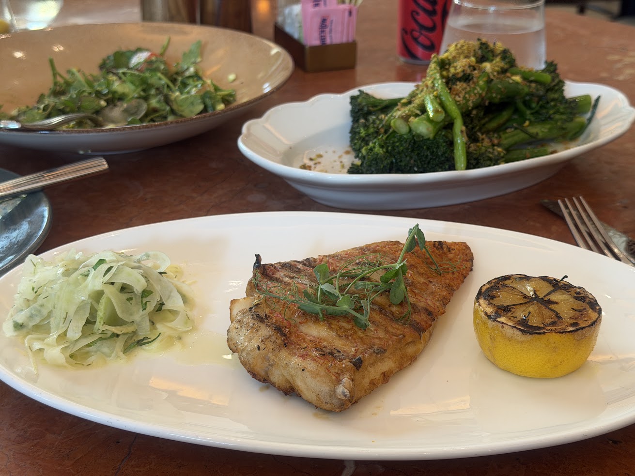 Grilled fish with herbs, a charred lemon, slaw, salad, and broccolini on a restaurant table—just one of the beautifully plated dishes you might enjoy while reading a Gale Miami Hotel & Residences Review.