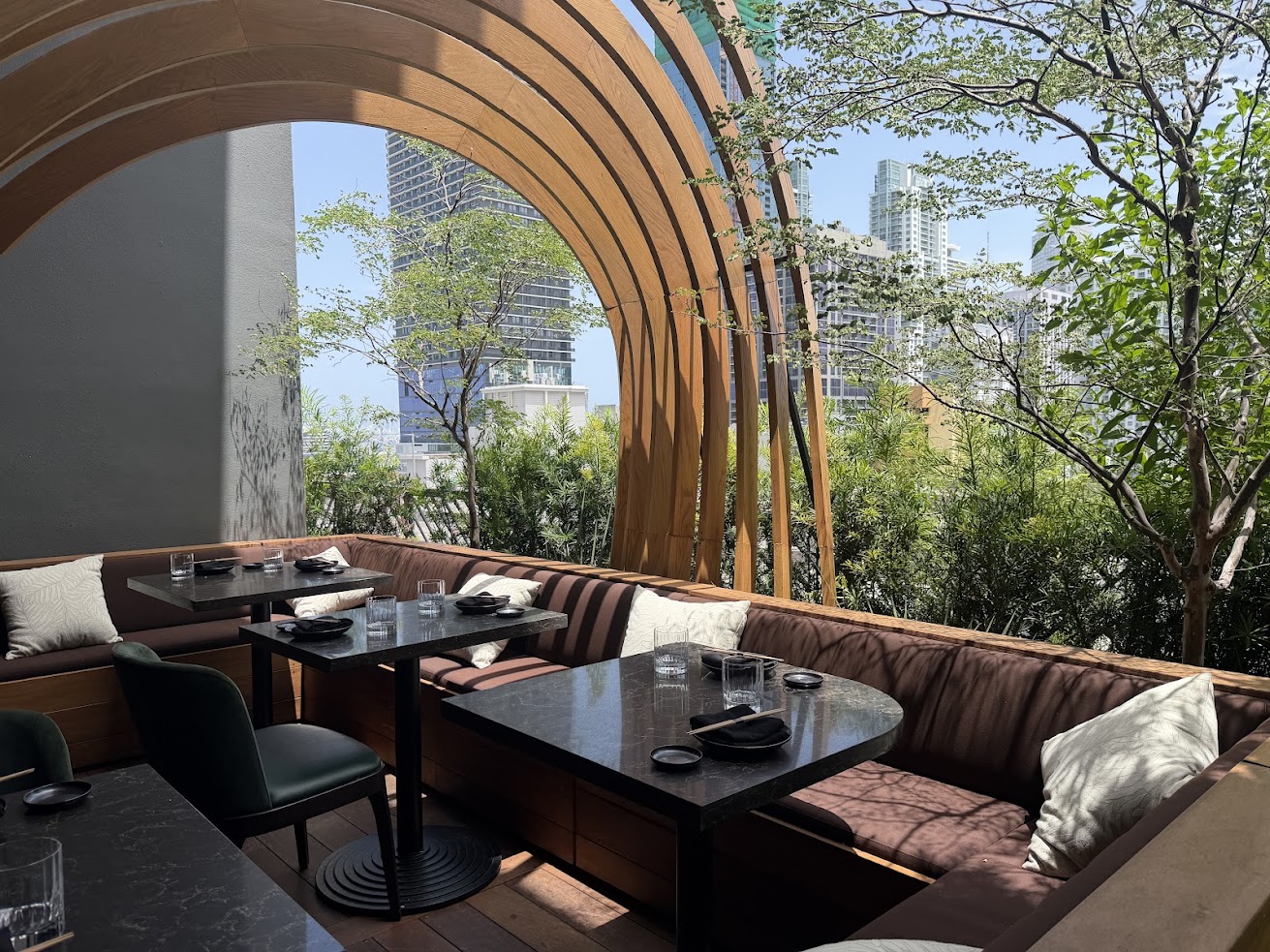 Outdoor restaurant seating with wooden arches, brown cushions, and city buildings visible through greenery creates a charming atmosphere reminiscent of the inviting spaces highlighted in any Gale Miami Hotel & Residences review.