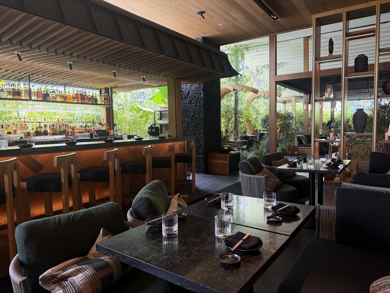 Modern restaurant interior with a bar, wooden furniture, glassware, and lush greenery outside large windows—creating an inviting atmosphere reminiscent of those highlighted in any thoughtful Gale Miami Hotel & Residences Review.