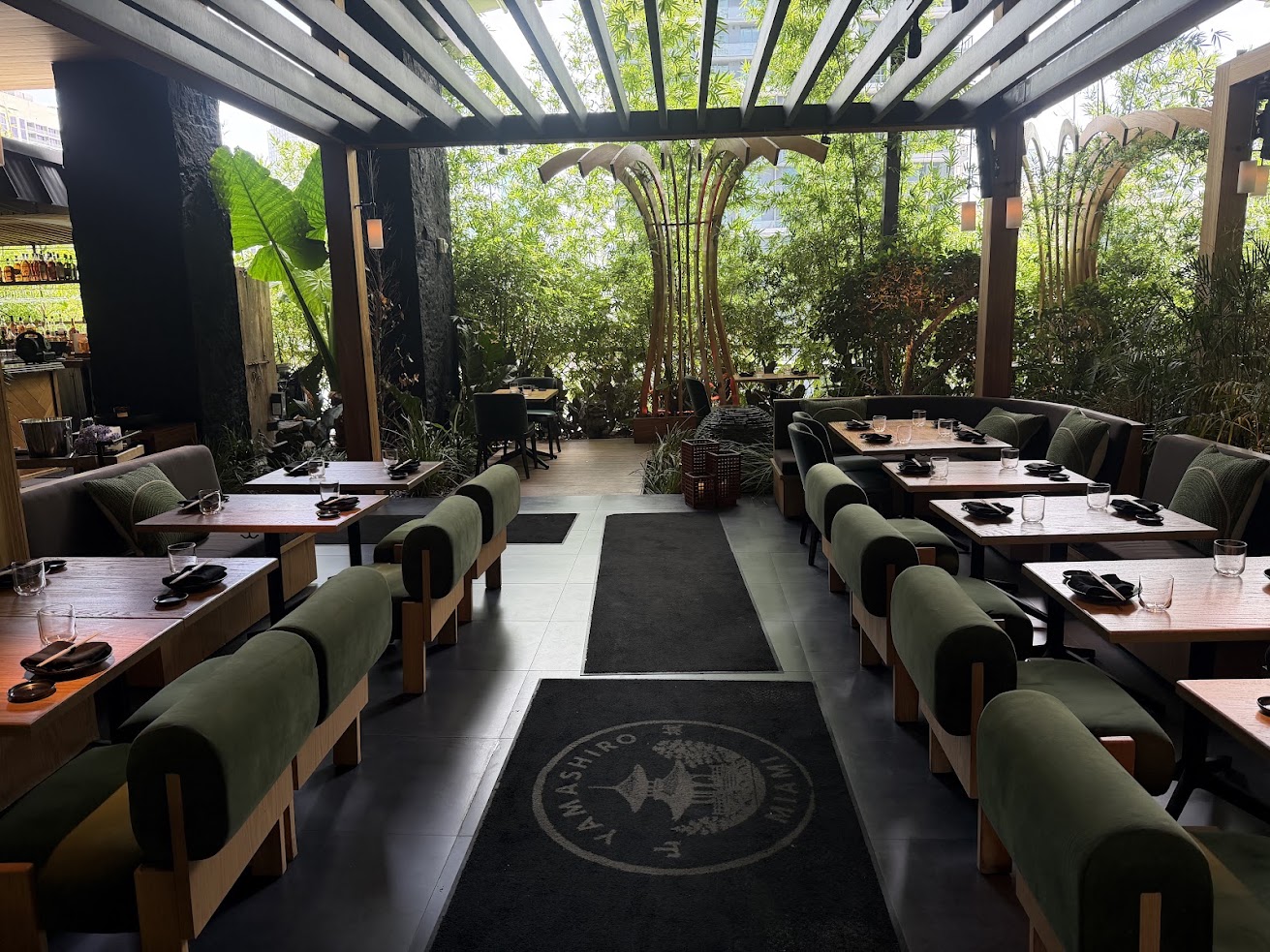 Modern restaurant with green cushioned seating, wooden tables, and lush greenery visible through large windows—a setting reminiscent of the stylish ambiance described in many Gale Miami Hotel & Residences reviews.
