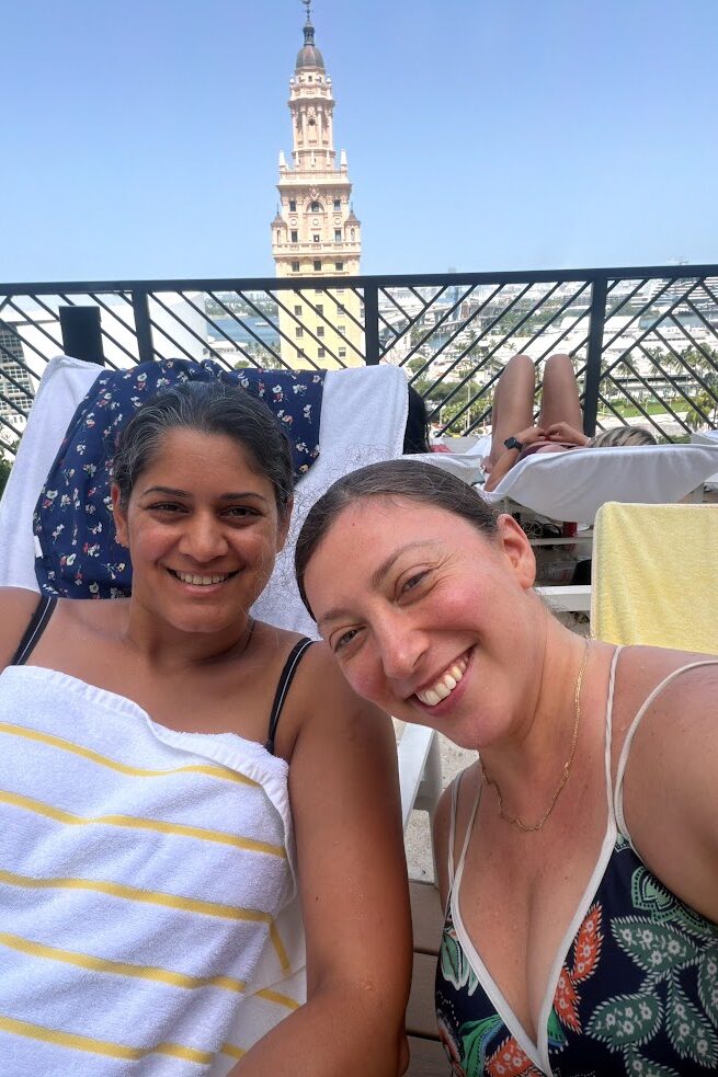 Two women smiling by a pool at the Gale Miami Hotel & Residences, with a tall building and blue sky in the background, capturing a perfect moment for this Gale Miami Hotel & Residences review.