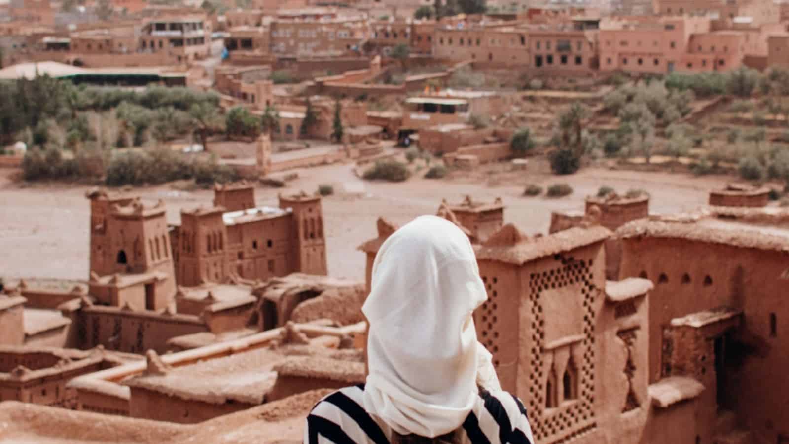 A person with a white headscarf overlooks an ancient, brown clay city with buildings and greenery in the distance.