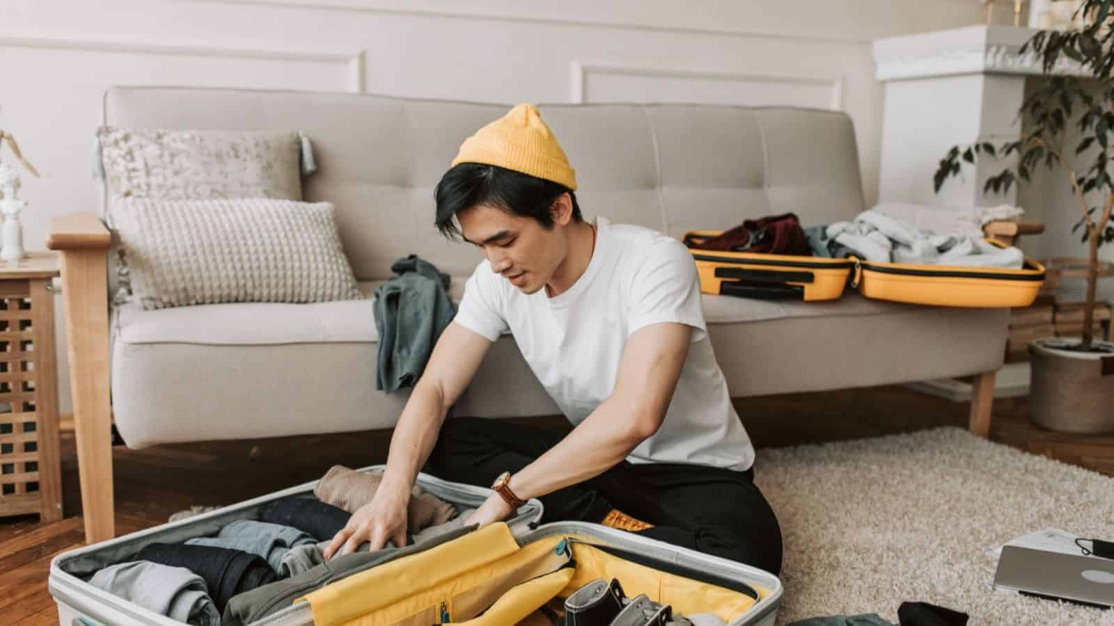 Man in a yellow beanie packing clothes for cold weather into an open suitcase in a living room with a sofa behind him.