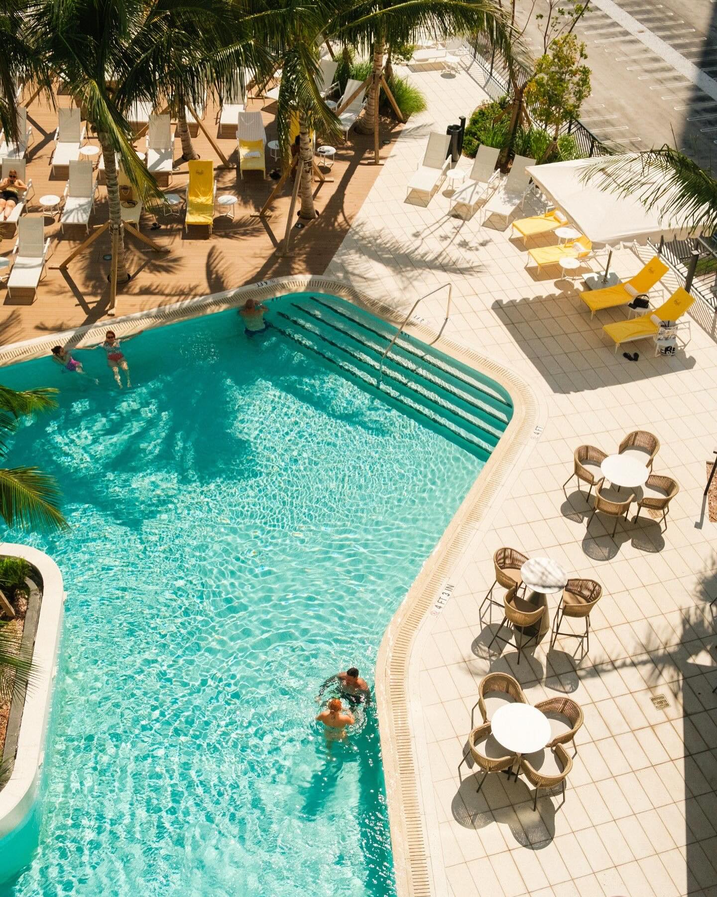 Aerial view of a sunny outdoor pool at the Gale Miami Hotel & Residences, with people swimming, surrounded by lounge chairs, tables, and palm trees—perfect for a relaxing day in Miami.