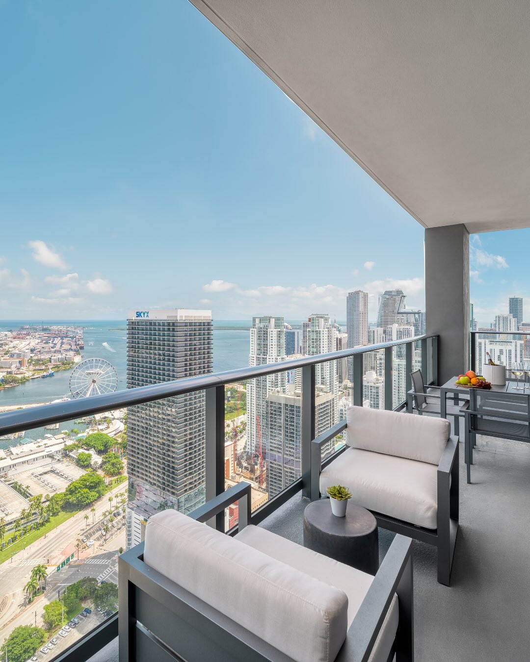 High-rise balcony with chairs, table, glass railing, and a city and waterfront view under a blue sky—perfect for experiencing the vibrant atmosphere highlighted in any Gale Miami Hotel & Residences review.
