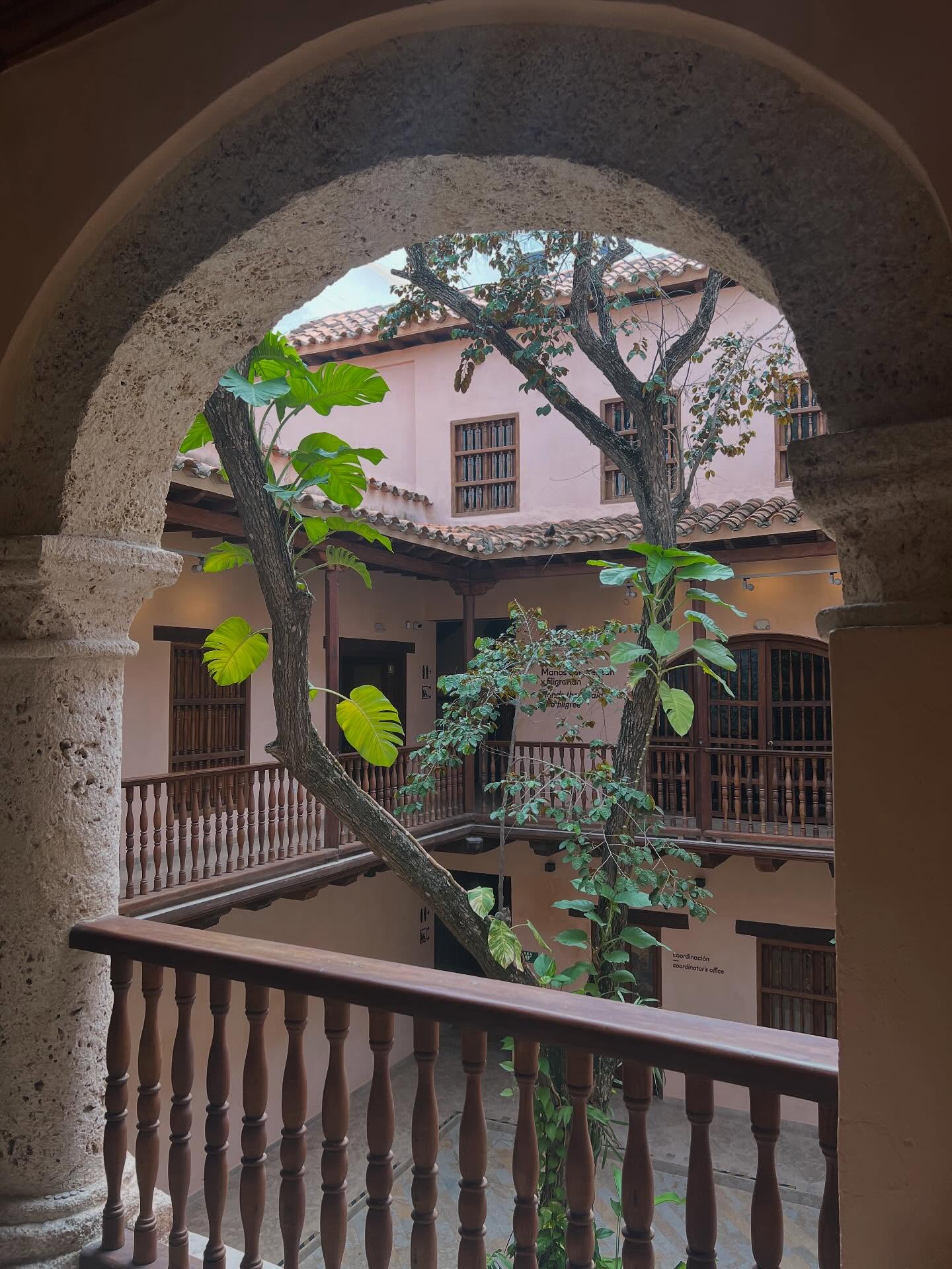 Stone arch frames a courtyard with leafy trees and pink walls, wooden railings, and barred windows&mdash;a hidden gem that captures the charm of old town and is among the many things to do in Cartagena.