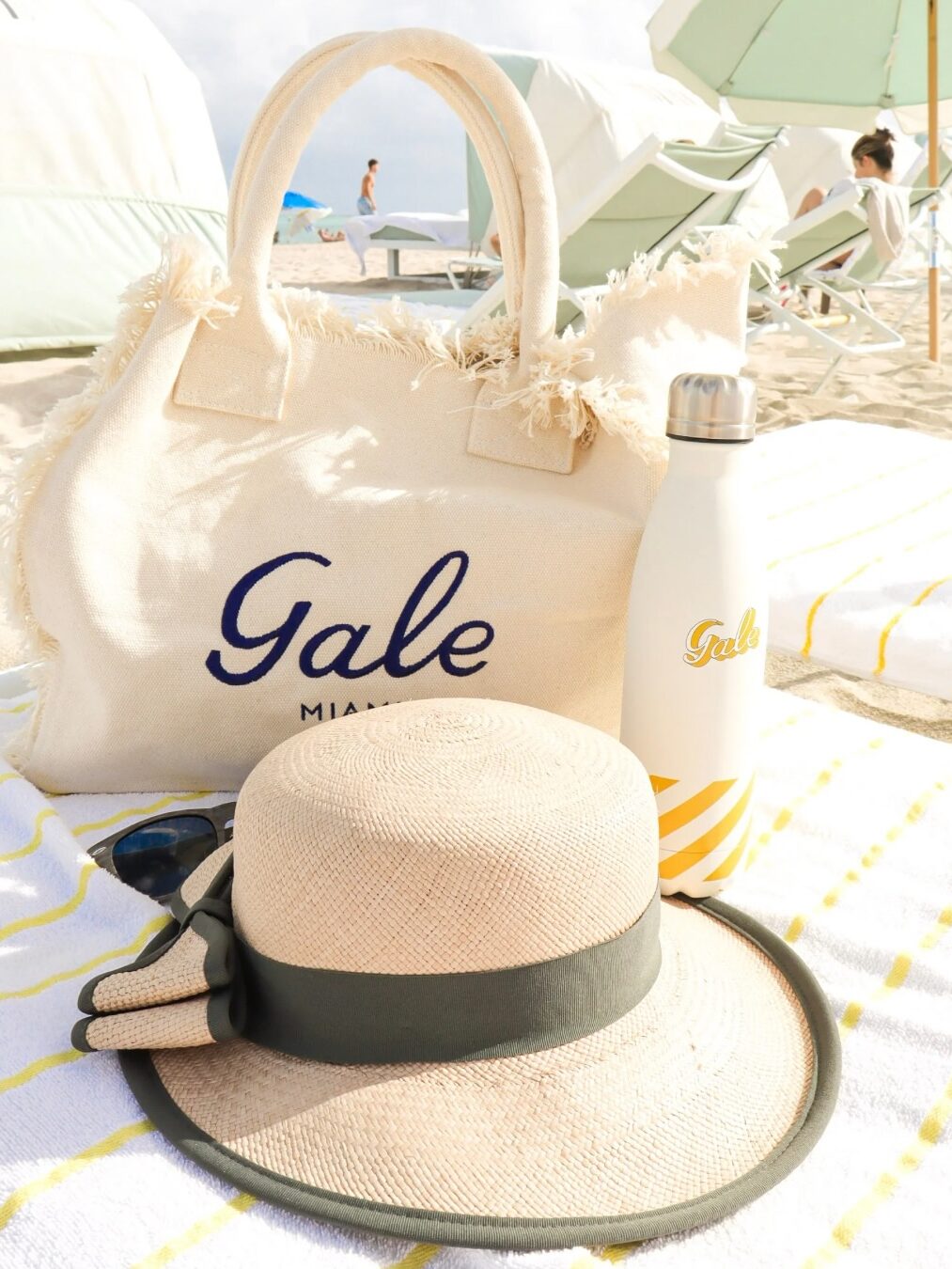 Beige hat, tote bag, and water bottle on a striped towel at a sunny beach with umbrellas—capturing the perfect moment of relaxation inspired by our recent Gale Miami Hotel & Residences review.