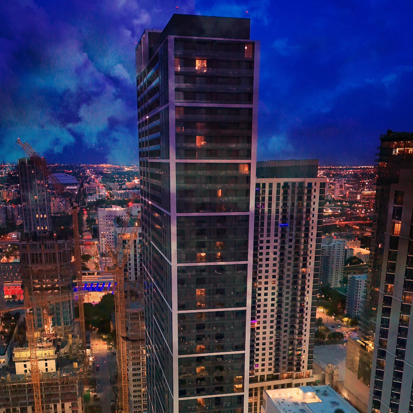Tall city buildings lit up at dusk, with construction cranes and a dark blue, cloudy sky in the background—a scene reminiscent of the skyline featured in many a Gale Miami Hotel & Residences review.