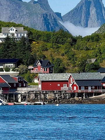 Red houses by the water with boats, green hills, and mountains in the background under a blue sky.