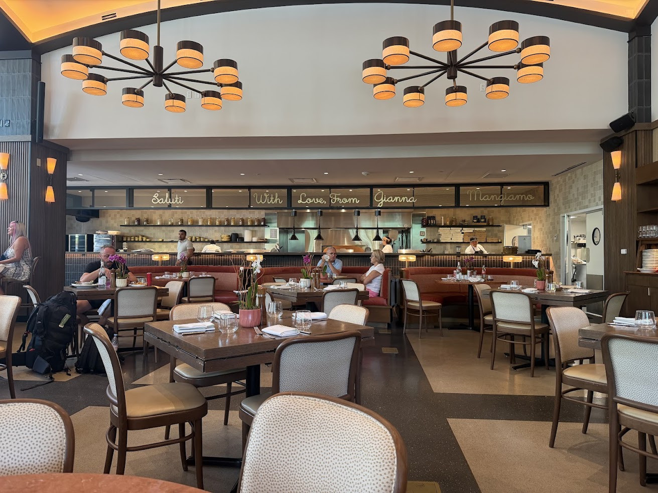 Restaurant interior with tables, people dining, open kitchen, and a sign reading “Saluti With Love, From Gianna Mangiamo”—a setting reminiscent of the sophisticated ambiance noted in the Gale Miami Hotel & Residences Review.