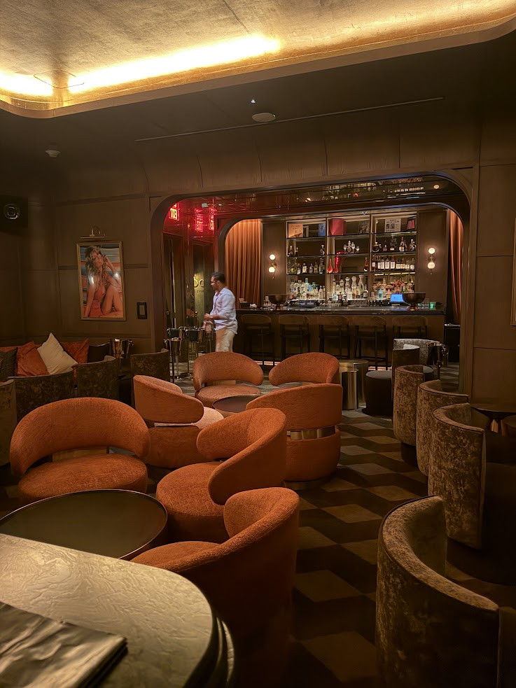 A lounge bar with orange chairs, a stocked bar, dim lights, and a person standing by the counter—just one of the inviting spaces you might discover in a Gale Miami Hotel & Residences Review.