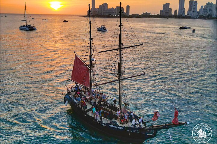 A pirate-style ship with red sails sails at sunset near a city skyline, crowded with people&mdash;a picturesque scene that highlights the unique and vibrant things to do in Cartagena.