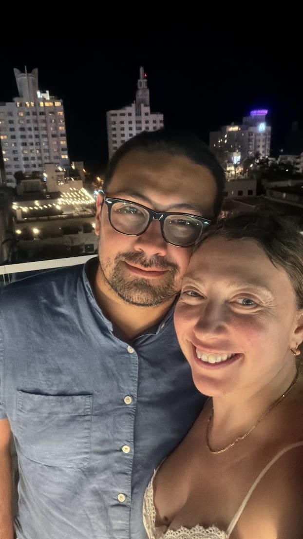 A man and woman smiling for a selfie at night with a Seia Miami Review city skyline in the background.