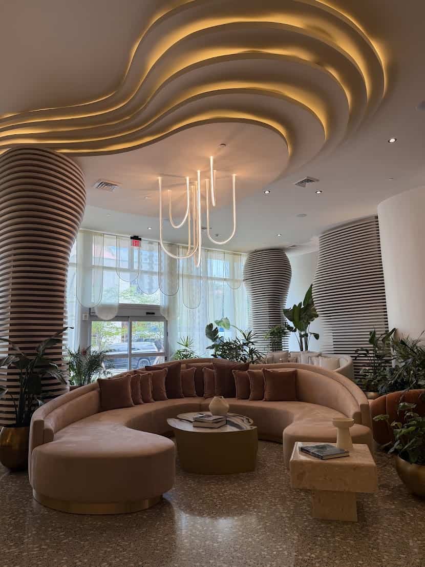 A curved, blush-pink velvet sofa in a modern Seia Miami lounge with illuminated ceiling panels and ribbed columns.