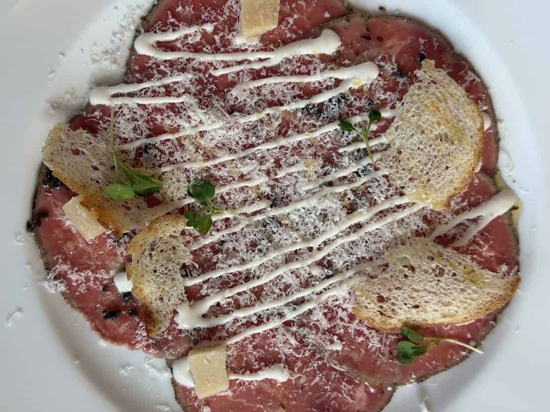 Beef carpaccio from a Seia Miami review, topped with grated cheese, a creamy drizzle, croutons, and microgreens.