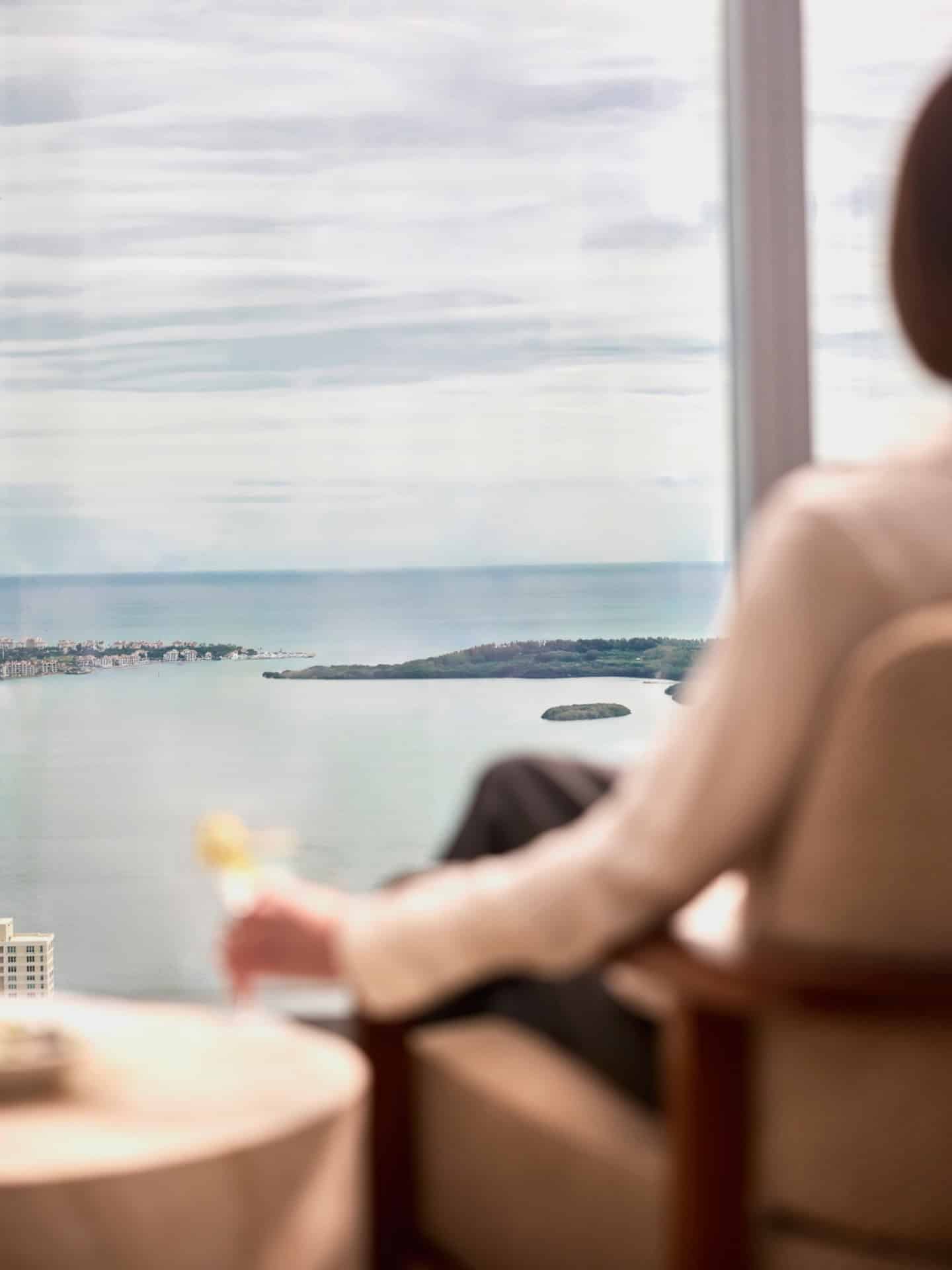 A person sits in a chair by a large window overlooking a bay, as seen in our Seia Miami review.