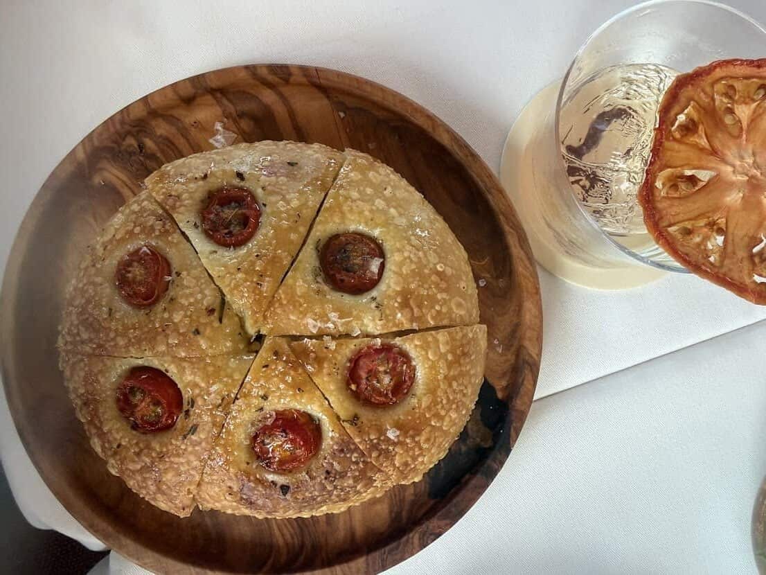 Round focaccia with cherry tomatoes on a wooden plate next to a citrus cocktail, as seen in a Seia Miami review.