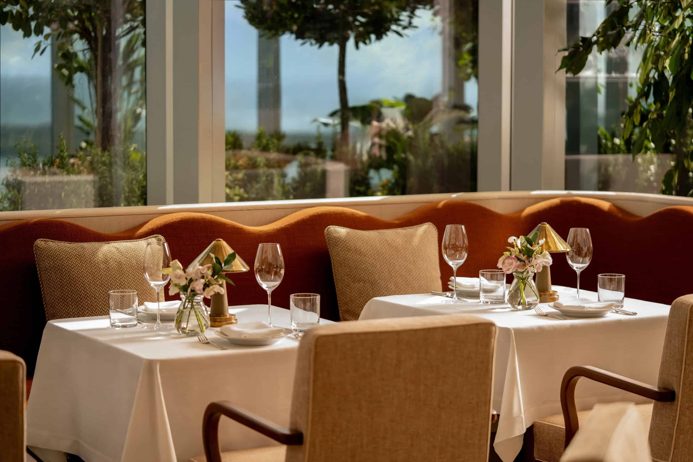 Two tables with white tablecloths, wine glasses, and flower vases at Seia Miami with large windows.
